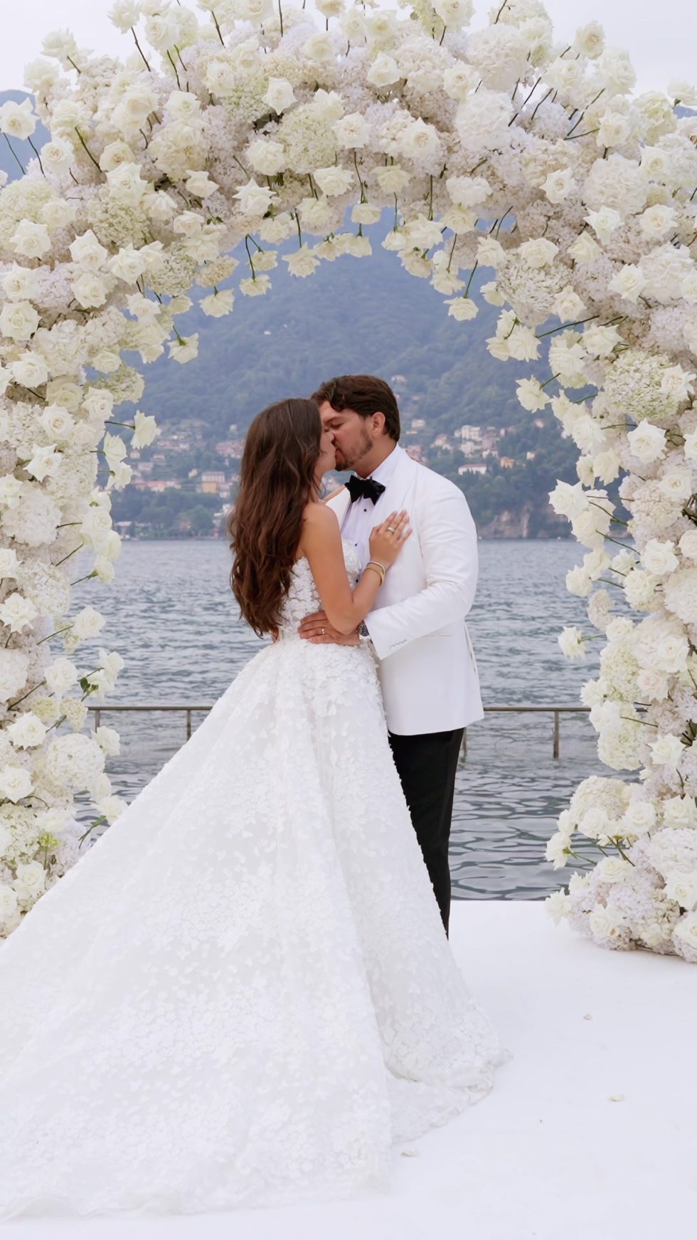 Renee and Tino embracing beneath the floral arch on Lake Como