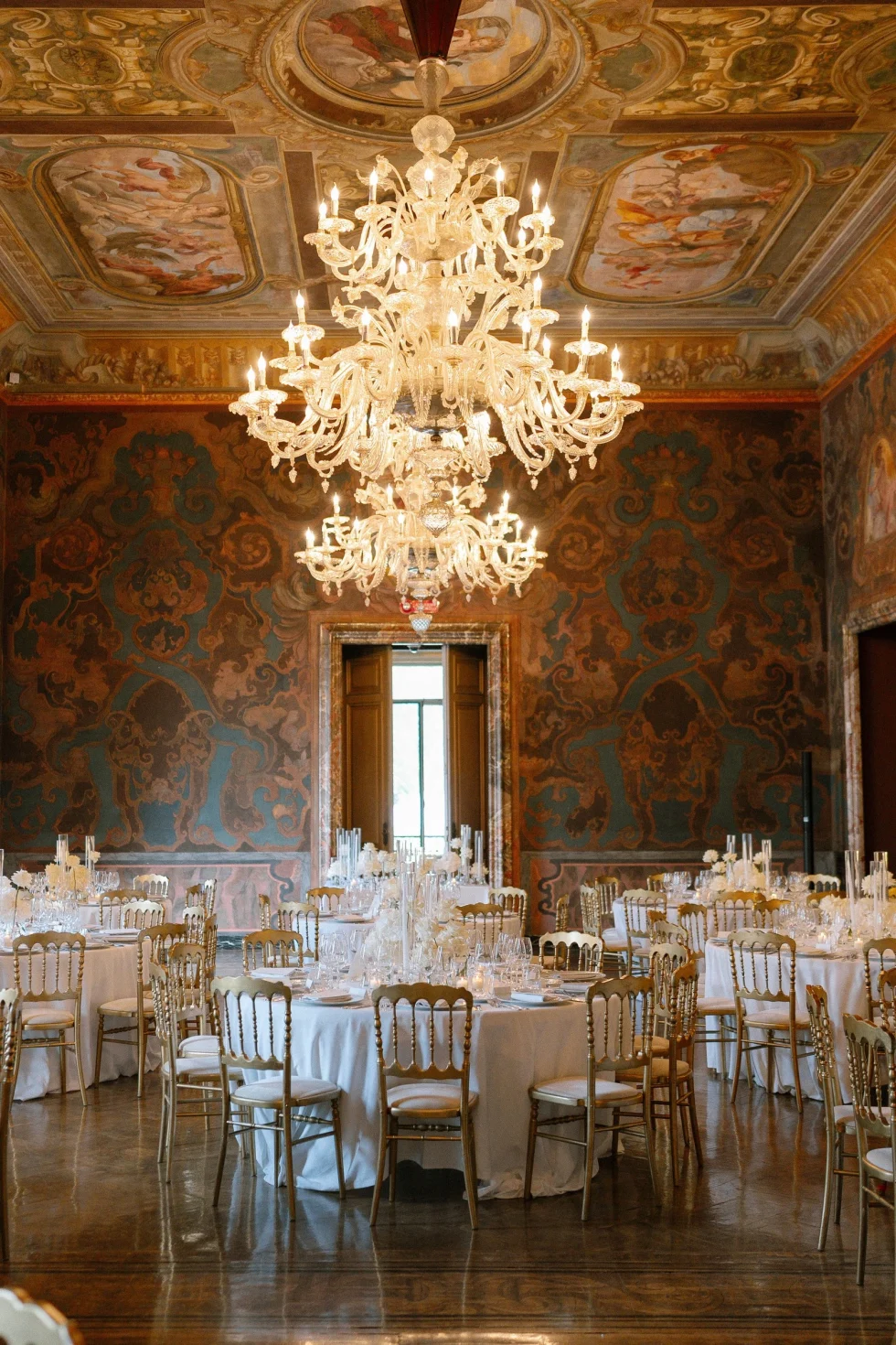 Wedding dinner set beneath Murano chandeliers in a frescoed hall of Villa Erba, Lake Como
