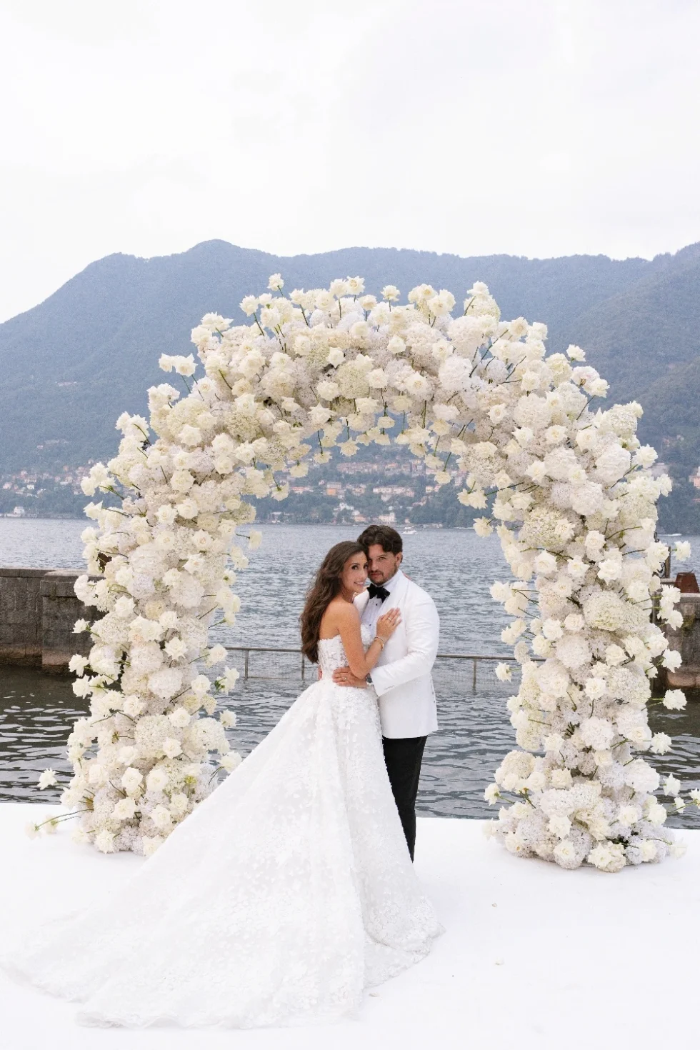 Renee and Tino embracing beneath the white rose arch on Lake Como at Villa Erba